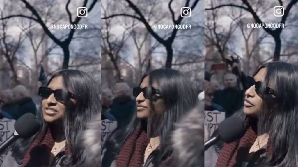 A young woman at an outdoor protest rally speaks seriously to an off-camera interviewer while holding a sign, unaware she is responding earnestly to a pun-based trick question about the Strait of Hormuz.