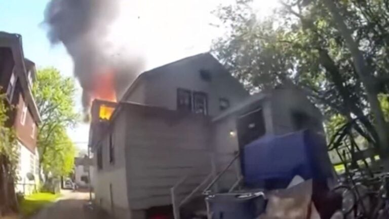 A bodycam screenshot showing a Michigan public safety officer positioned near a second-floor window of a burning house as smoke pours out above him.
