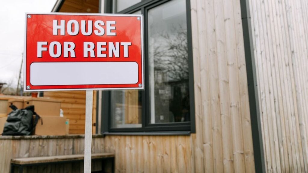 Red 'House for Rent' sign outside modern wooden house.