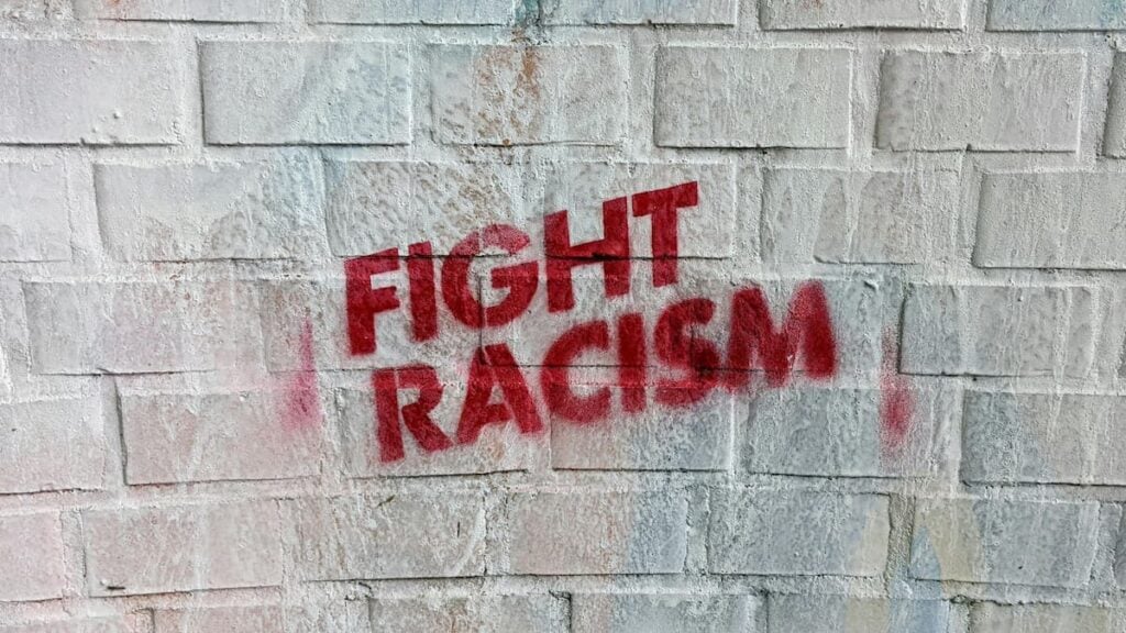 Texas Woman Asks Whether Black Community Is Backing Beauty Store Boycott, Debate Quickly Erupts: ‘If You Have To Ask That, You’re Not Serious About It!’ Red spray-painted graffiti reading “Fight Racism” on a white brick wall