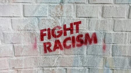 Texas Woman Asks Whether Black Community Is Backing Beauty Store Boycott, Debate Quickly Erupts: ‘If You Have To Ask That, You’re Not Serious About It!’ Red spray-painted graffiti reading “Fight Racism” on a white brick wall