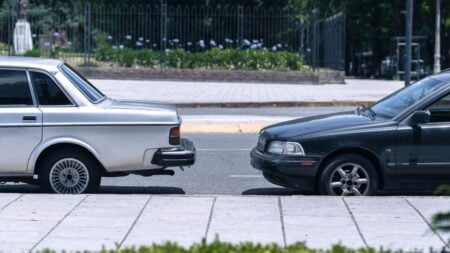 Two vintage cars parked on a sunny urban street amidst greenery and sidewalks.