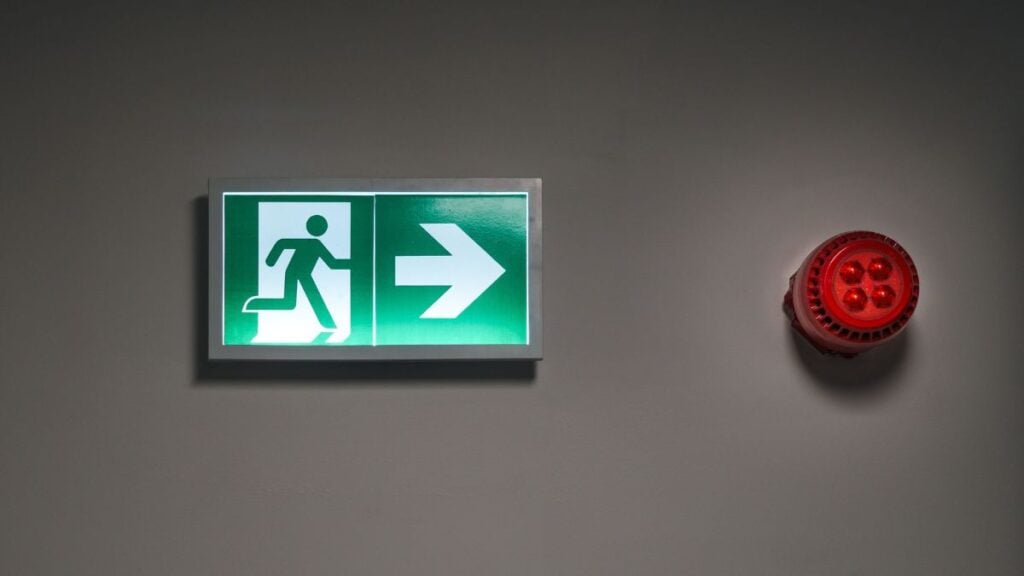Shirtless Ohio Resident Confronts Maintenance Worker Replacing Smoke Alarm, Threatens to Call ICE: ‘Go Ahead’ A brightly lit exit sign and alarm in an indoor corridor, indicating emergency escape route.