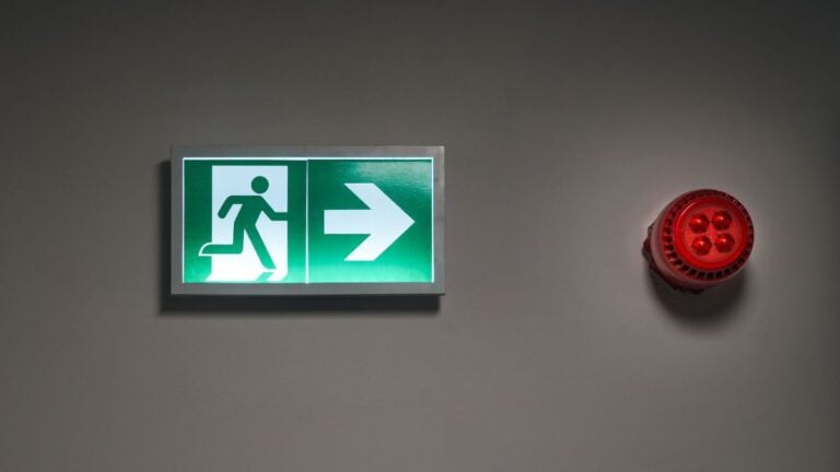 Shirtless Ohio Resident Confronts Maintenance Worker Replacing Smoke Alarm, Threatens to Call ICE: ‘Go Ahead’ A brightly lit exit sign and alarm in an indoor corridor, indicating emergency escape route.