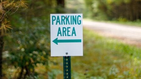 California Karen Chases Man Down Street After He Takes Parking Spot She Wanted: ‘Going Out Of Her Way To Harass People’ Close-up of a parking area sign with green text on a forest pathway.