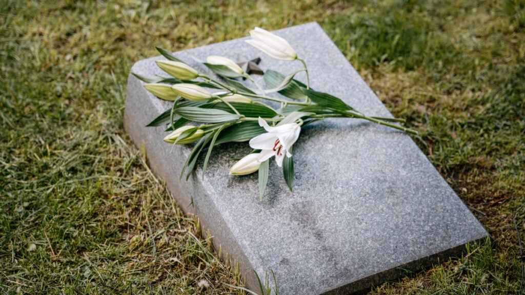 Shocked Oregon Resident Says They Found Their Own Obituary Online: ‘Sounds Like the Start to a Psychological Horror Movie’ White lilies placed on a gravestone in a cemetery