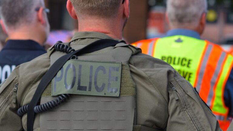Arizona Police Tackle Black Woman Onto Metal Bike After She Approaches Arrest Scene in Tempe: ‘This Is False Imprisonment and Harassment’ Rear view of police officers in tactical gear and high-visibility vests in a public setting.