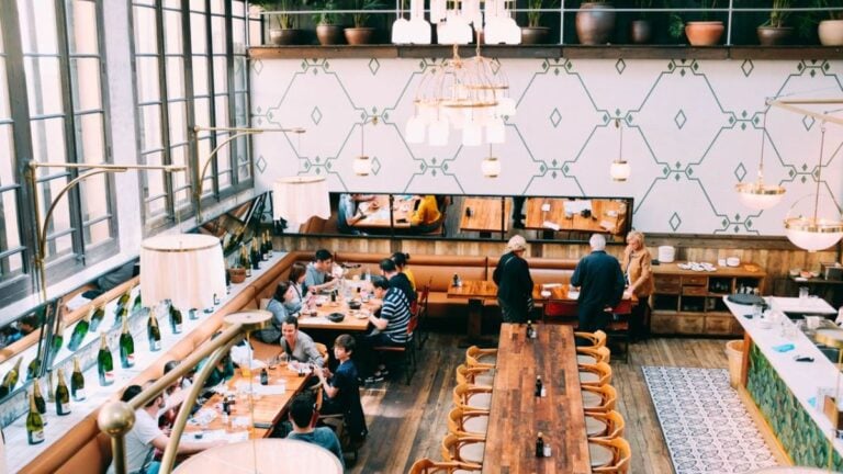 Black Woman Accuses Florida Restaurant Of Racism After Staff Allegedly Locked Doors Thinking She Wouldn’t Pay: ‘The Treatment Is Disgraceful’ Spacious restaurant with stylish interior design, featuring wooden tables and a group of diners enjoying a meal.