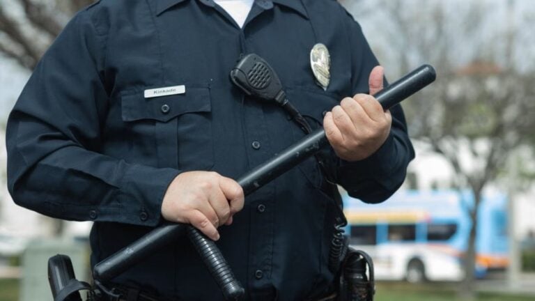 California Woman Calls Officers ‘Human Traffickers’ While Accusing Them Of Assisting ICE: ‘Karens Are All The Same’ Close-up of a police officer holding a baton outdoors, emphasizing authority and security.