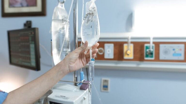 North Carolina Man Accuses Hospital of Racism, Refuses Medical Care While Officers Stand Nearby: ‘I Don’t Want to Be Seen’ Close-up image of an IV drip and hand in a hospital room, illustrating medical care.