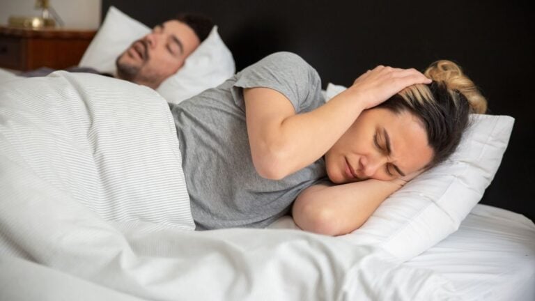 ‘How Does He Forget Something That Keeps Him From Dying?’: New Jersey Woman’s Vacation Marriage Dispute Goes Viral A woman looks frustrated in bed as her partner snores loudly, highlighting sleep disturbance issues.