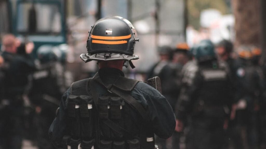 Police officer in full riot gear with blurred background of officers in action during daytime patrol.