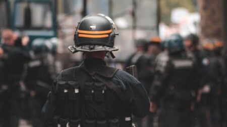 U.S. Marshals and Police Come Crashing Down as Deck Collapses During Search Warrant in Missouri: ‘It’s Raining Men’ Police officer in full riot gear with blurred background of officers in action during daytime patrol.