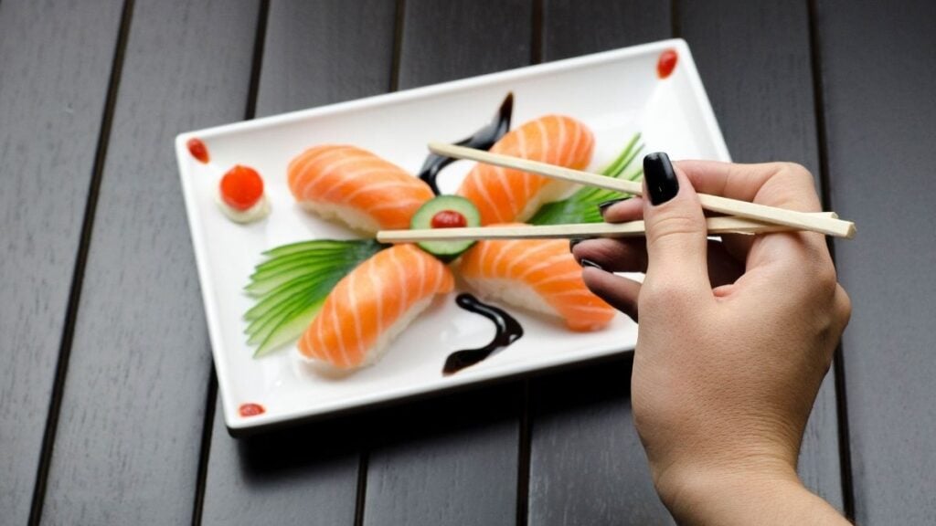 New York Sushi Customer Refuses to Pay Bill After Claiming Staff Laughed at Them: ‘I’m Not Paying for the Food’ Close-up of a sushi plate with salmon nigiri and decorative garnish, held with chopsticks.