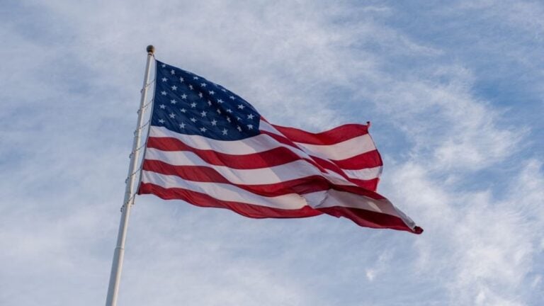 Oregon Woman Tries to Grab American Flag at Patriot Rally, Ends Up Crying on Grass: ‘She Is a Thief’ The American flag waves proudly against a backdrop of blue sky, symbolizing freedom and patriotism.