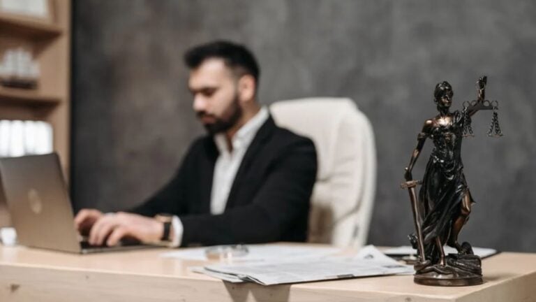 California Immigration Law Firm Accused of Threatening to Call ICE on Clients Who Wanted Their Money Back: ‘Seems Everyone Is Corrupt’ A businessman working on a laptop in an office with a Lady Justice statue in focus on the desk.