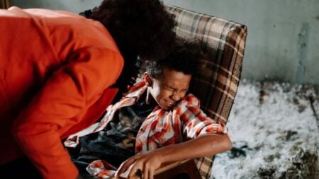 Child in distress on checkered chair with adult leaning over ominously inside a dimly lit room.