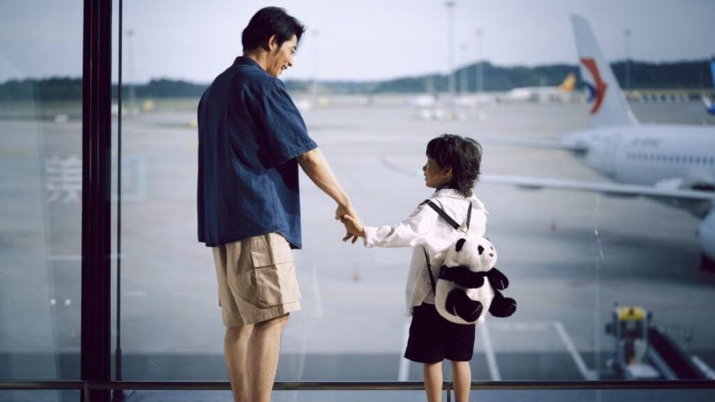 Hawaii Airport Staff Spot Young Child Wandering Around Baggage Drop-Off With No Parent in Sight: ‘That Kid Is Way Too Small to Be Exploring’ A father and child holding hands, gazing at planes through an airport window.