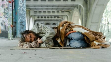 A homeless man lies under an urban bridge, wrapped in a blanket, symbolizing poverty and survival.