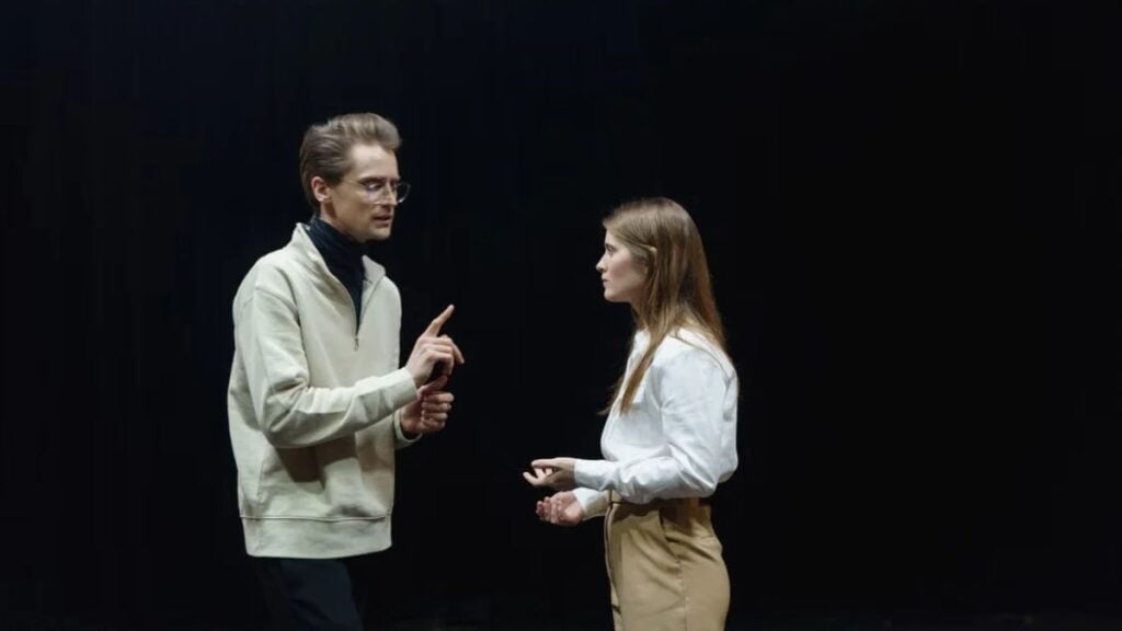 Texas High School Cancels Entire Musical After Girl Is Cast as Male Lead: ‘They’ve Never Heard of Shakespeare’ Two actors rehearsing on a dimly lit theater stage, focusing on performance and dialogue.