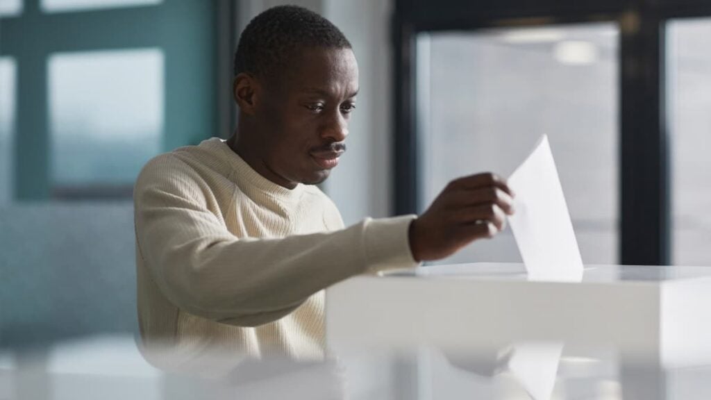 ‘They Want These Voices Silenced’: North Carolina Students March to Vote After Officials Deny Campus Polling Site A young man casting his vote at an indoor polling station, focused on the democratic process.